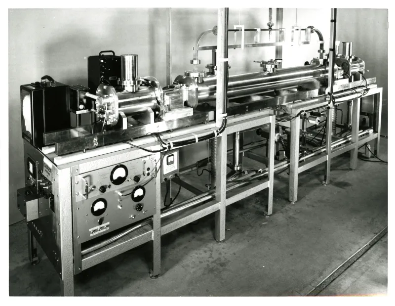 A black-and-white photograph of an early caesium beam frequency standard: a long horizontal vacuum tube of polished metal, flanged and clamped, running the length of a metal-framed bench, with a control panel of round dials and switches at the near end and cabling beneath