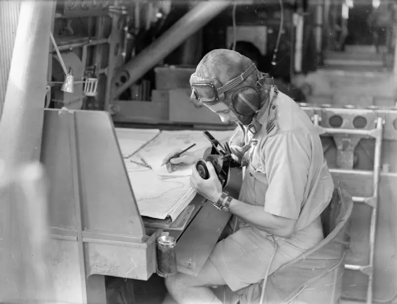 A black-and-white photograph inside an aircraft: a man in a flying helmet with goggles pushed up on his forehead, in short-sleeved shirt and shorts, sits at a chart table holding a bubble sextant to his eye with both hands, charts and a pencil on the table before him and the ribbed interior of the fuselage behind