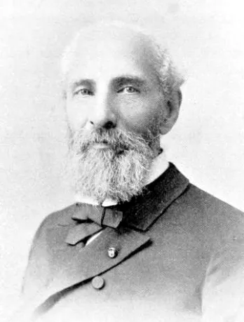 A photographic portrait of an elderly balding man with a full white beard and moustache, in a dark high-buttoned coat with a bow tie and a small lapel pin