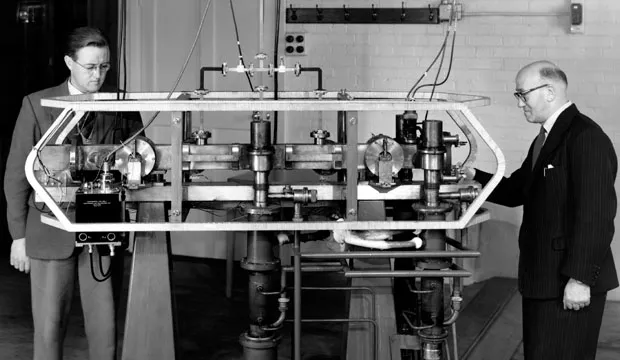 A black and white photograph of two men in suits standing at either end of a long laboratory apparatus on a bench: a horizontal metal framework carrying cylindrical vacuum components, valves, wiring and instrument boxes, against a painted brick wall
