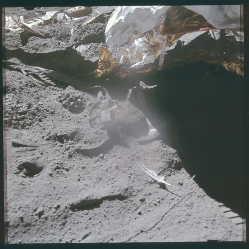 A square Hasselblad frame of grey lunar soil covered in bootprints. Gold and silver foil from the lunar module fills the upper right, with deep black shadow beside it. A metal geological hammer lies half-buried in the dust at the centre, one sunlit face bright. Below and to the right of it, a small pale feather rests on the surface. Cross-shaped register marks are scattered across the frame