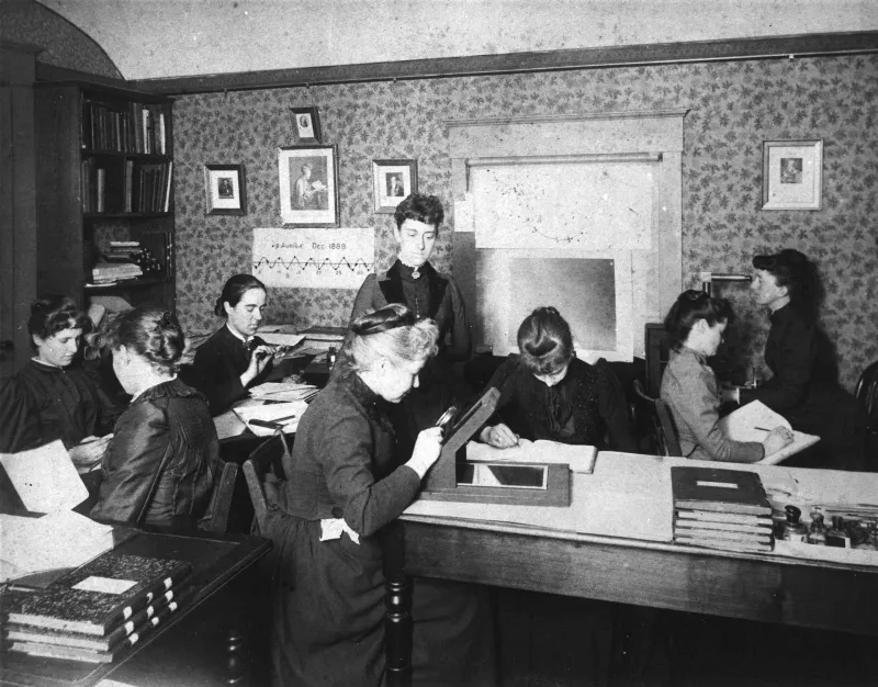 A photograph of eight women in dark high-necked dresses working in a wallpapered room: several seated at desks with papers and ledgers, one standing over a slanted desk examining a photographic plate through a magnifier on a light box, stacks of bound volumes and plate boxes on the tables, framed portraits and a chart on the walls
