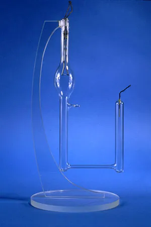 A museum object photograph against a deep blue ground: a slender bent glass tube rising from a clear circular base, carrying a small bulb near the top, with a separate short glass tube standing beside it
