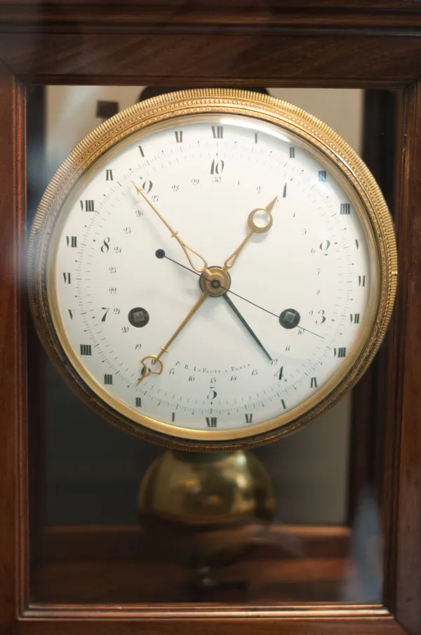 A mahogany-cased clock photographed through its glass door. The large white enamel dial sits in a gilt bezel and carries Arabic numerals 1 to 10 as its principal hours, with small Roman numerals I to XII around the outer rim and a finely divided minute track outside those. It is signed P. B. Lepaute a Paris, and a brass pendulum bob hangs below the dial