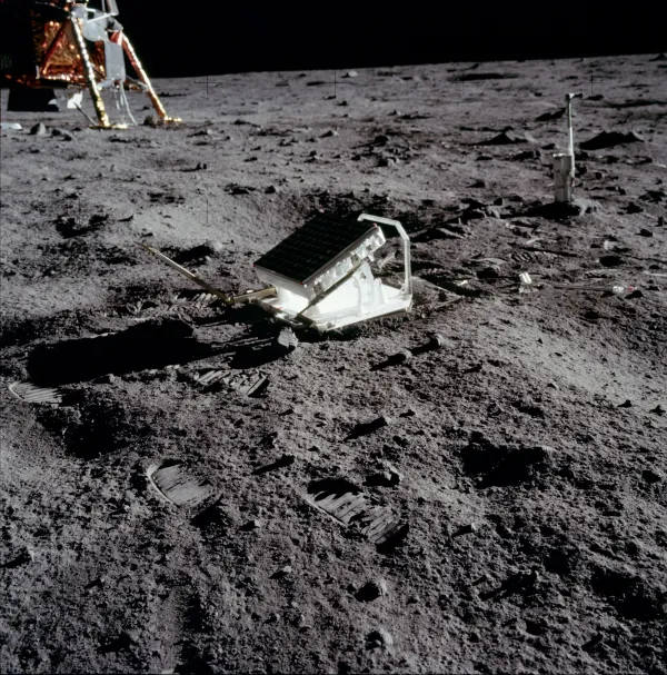 A photograph on the lunar surface: a tilted panel of small square corner-cube reflectors in a white frame, standing on grey dust scattered with boot prints, with a leg and gold foil of the Lunar Module at the upper left, a camera on a stand at the right, and black sky above the horizon