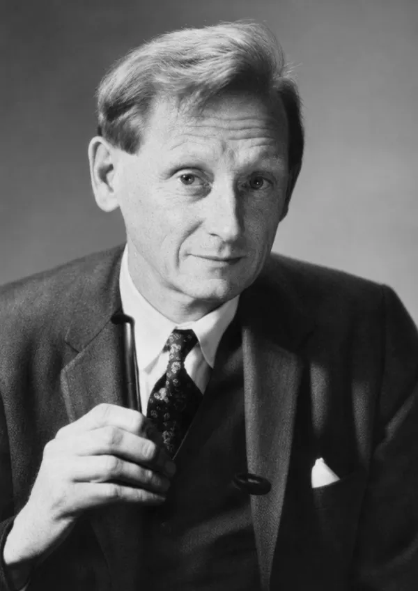 A black-and-white studio portrait of an elderly man in three-quarter view against a plain grey ground. He has swept-back greying hair and a lined face, and wears a dark tweed jacket over a white shirt with a dark patterned tie and a white pocket square. He holds a pipe upright in his right hand at chest height, and looks towards the camera with a slight smile