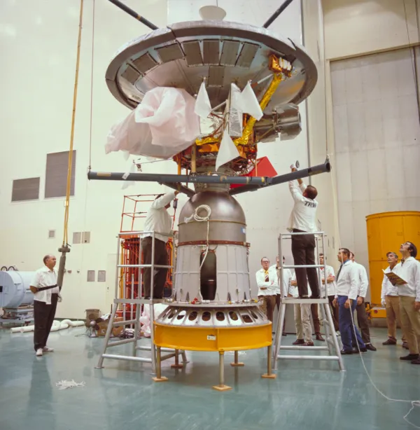 A colour photograph in a clean high-bay: a spacecraft hanging above a conical kick motor on a yellow stand, its large dish antenna uppermost, instrument booms folded against the body, two technicians in white coats on stepladders working on it and several engineers in shirtsleeves standing about with clipboards