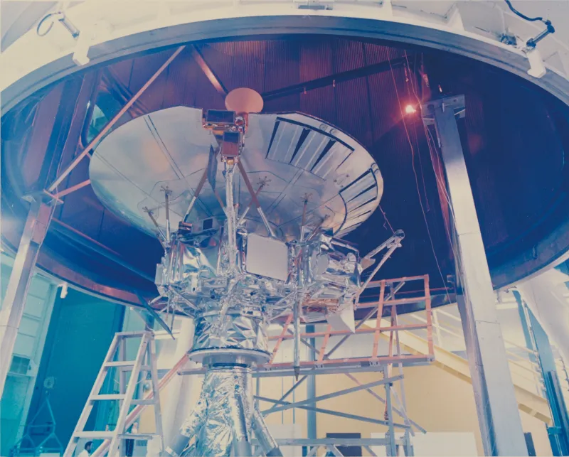 A faded colour photograph from 1971: a spacecraft wrapped in crinkled silver multilayer insulation standing on a stand inside a large test chamber, its parabolic dish above it, the dark ribbed wall of the chamber shroud curving behind, with scaffolding, a stepladder and a single red-glowing lamp high on the right
