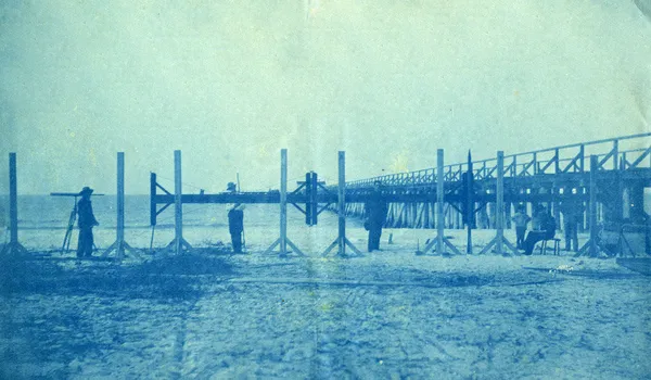 A blue-toned cyanotype photograph of a flat sandy beach with calm water to the left. A long low wooden trestle runs away from the foreground into the distance, supported on posts driven into the sand, with several men standing along it; one at the left works at a tripod-mounted instrument