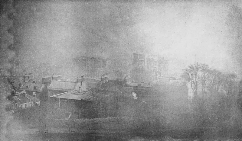 A faded, fogged monochrome photograph of a hillside town: slate rooftops, chimneys and dormers in the foreground, a terrace of houses at the lower left, and in the upper right a group of tall buildings partly veiled in mist, one of them covered in a lattice of scaffolding. Bare leafless trees stand at the right of the frame