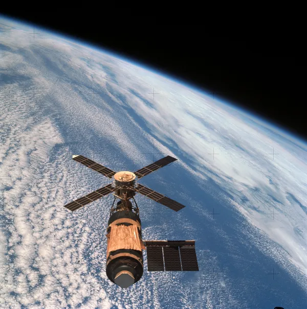 Looking down on Skylab against the Earth: a four-armed windmill of solar panels around a gold-brown telescope mount at the top, a cylindrical workshop below it draped in gold thermal material, a single rectangular solar wing extending to one side, and the docking end pointing towards the camera. Beneath is a field of white cloud over blue ocean, and along the top the atmosphere shows as a thin bright band against black space. Small cross-shaped register marks from the camera are scattered across the frame