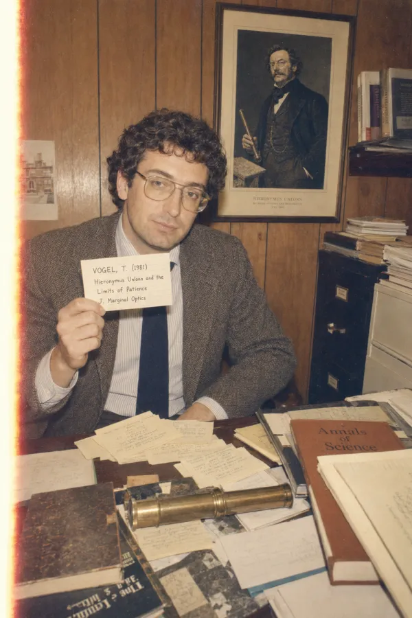 Faded 1981 colour photograph of a tweed-jacketed academic at a cluttered desk, holding a single index card and looking patiently at the camera