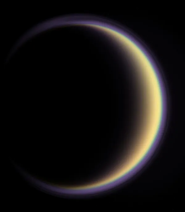 Titan as a thin crescent: a dark globe with a golden sunlit limb at the right, encircled by a complete violet halo of haze that continues around the unlit side