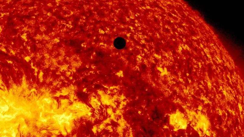 Photograph of the Sun's chromosphere in deep red and orange, densely mottled and flecked with bright yellow, the limb curving away into black at the upper right, a bright active region at the lower left, and Venus crossing as a small, perfectly circular, wholly black disc